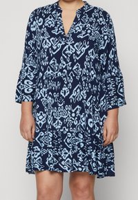 Kvinna iklädd en löst sittande marinblå klänning med ljusblå abstrakta mönster och trekvartsärmar, stående mot en enfärgad bakgrund.