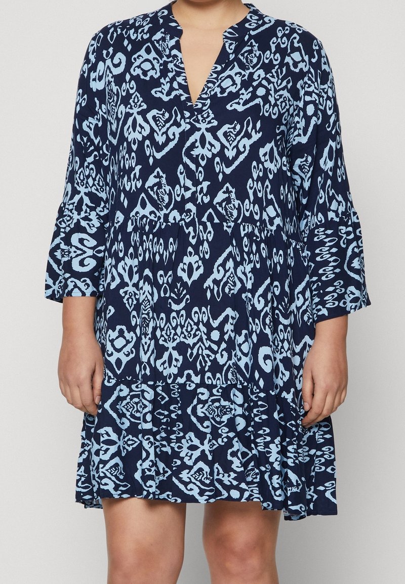 Kvinna iklädd en löst sittande marinblå klänning med ljusblå abstrakta mönster och trekvartsärmar, stående mot en enfärgad bakgrund.