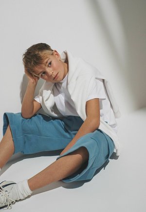 Junge mit kurzen Haaren sitzt auf dem Boden, trägt ein weißes T-Shirt, blaue knielange Shorts, weiße Turnschuhe und einen weißen Pullover, der über die Schultern gelegt ist.