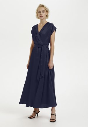Marinblå om_wrap-klänning med korta ärmar, djup V-hals och ett matchande bälte; flödande tyg med en mjuk textur ner till anklarna. Svarta sandaler.