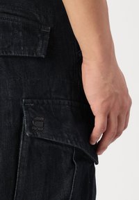 Black denim pants featuring a side pocket, a small embossed logo, and a textured fabric. Casual design with a relaxed fit.