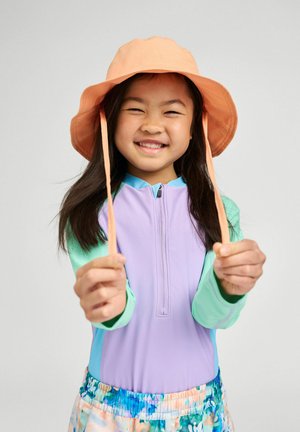 Uśmiechnięta dziewczynka w brzoskwiniowym kapeluszu przeciwsłonecznym i kolorowym kostiumie kąpielowym z długim rękawem trzyma sznurki kapelusza do przodu na jednolitym tle.