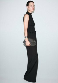 Donna in un vestito nero senza maniche che tiene una borsa a clutch texture, indossando bracciali d'argento, in piedi di lato contro uno sfondo chiaro e uniforme.