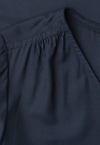 Tissu bleu foncé avec un détail de couture froncée près d'une couture et de l'encolure, montrant la texture et la construction d'un vêtement.
