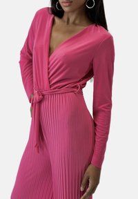 Jumpsuit scollato a V profondo in un brillante colore rosa, con maniche lunghe, vita con cintura e texture plissettata nella parte inferiore.