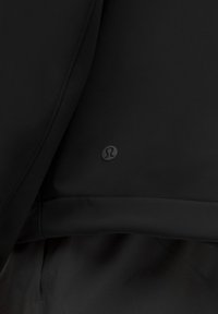 Juodas sportinis švarkas su lygiu audiniu, minimalistiniu dizainu ir mažo logotipo detale šalia apvadų. Nėra jokių kitų pastebimų raštų ar akcentų.
