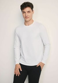 Biała koszulka z długim rękawem i okrągłym dekoltem, wykonana z miękkiego materiału, z małym logo na piersi, zestawiona z czarnymi spodniami.