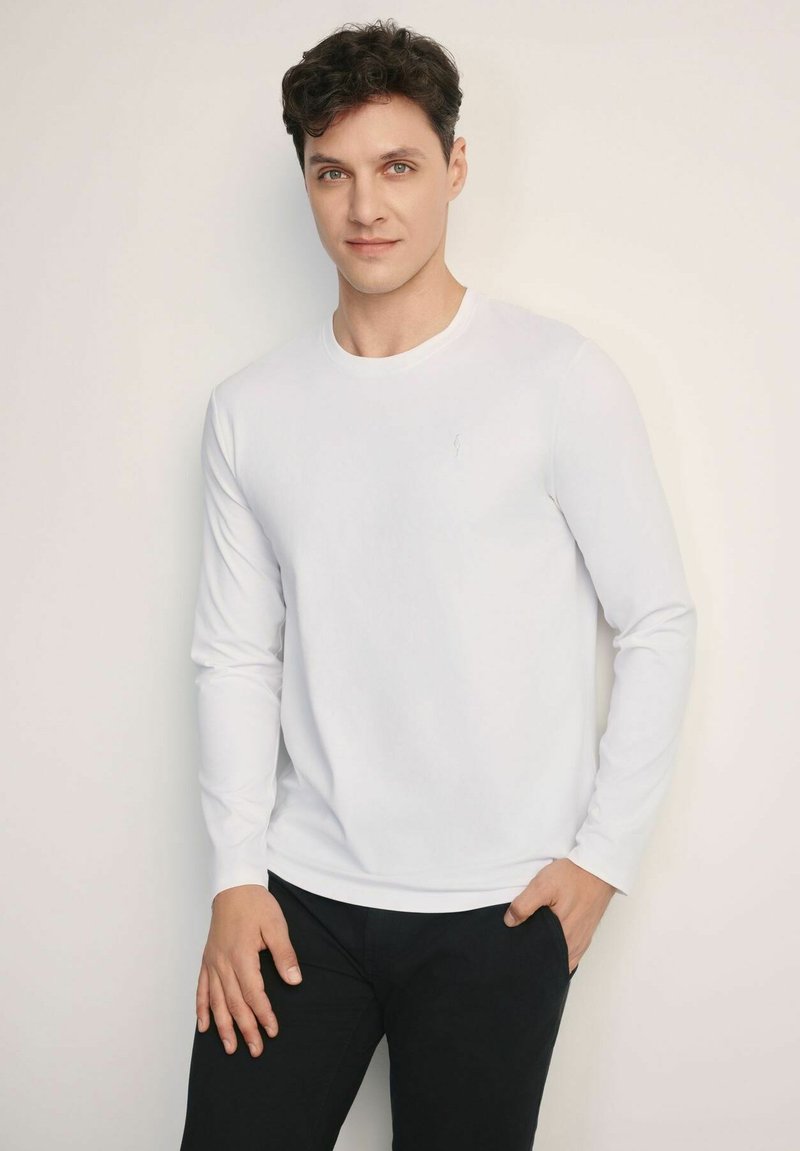 Biała koszulka z długim rękawem i okrągłym dekoltem, wykonana z miękkiego materiału, z małym logo na piersi, zestawiona z czarnymi spodniami.