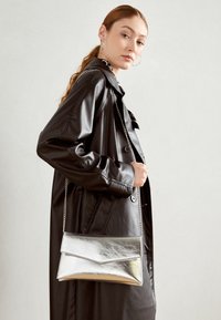 Metalická stříbrná kabelka na skladování s přeloženým víkem a řetízkovým popruhem, kterou drží modelka v černém koženém kabátu. Hladký povrch.
