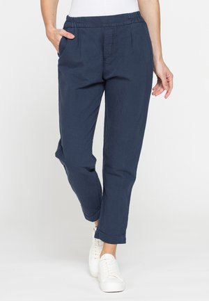 Persona che indossa pantaloni elasticizzati blu navy con orli risvoltati e sneakers bianche, una mano in tasca e cammina in avanti.