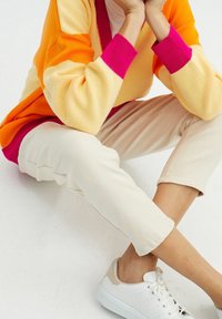 Sweter kardigan na drutach w odcieniach pomarańczowego i żółtego z różowymi mankietami, zestawiony z kremowymi zwężanymi spodniami i białymi trampkami z beżowymi akcentami.