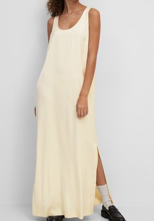 Femme portant une longue robe crème sans manches avec une fente latérale, associée à des chaussettes blanches et des mocassins noirs, debout devant un fond uni.