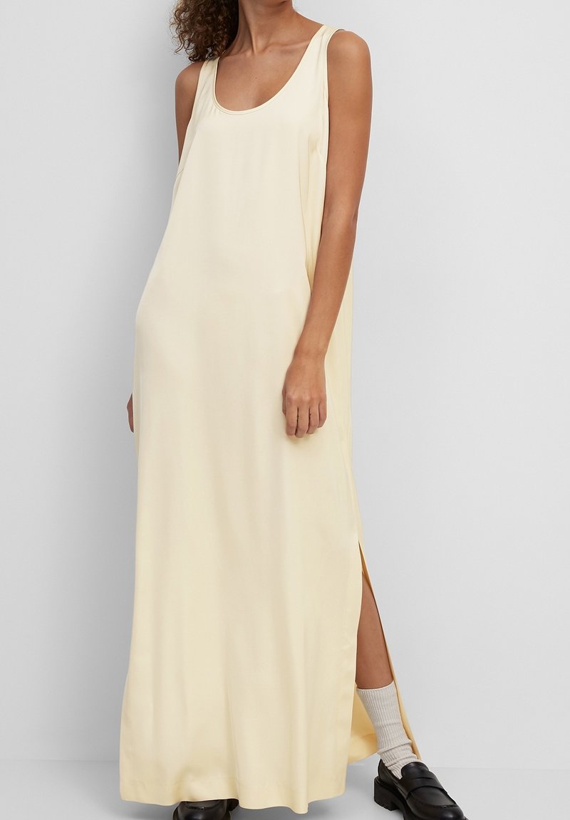 Femme portant une longue robe crème sans manches avec une fente latérale, associée à des chaussettes blanches et des mocassins noirs, debout devant un fond uni.