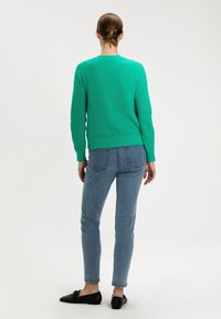 Zelený žebrovaný svetr s dlouhými rukávy, kombinovaný se světle modrými slim-fit džíny a černými mokasínami, viděný ze zadní strany.