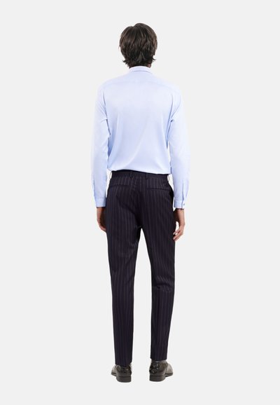 Himmelblaues Langarmshirt mit taillierter Passform; kombiniert mit dunkelblauen, gestreiften Hosen. Das Model steht mit den Händen an den Seiten und trägt schwarze Schuhe.