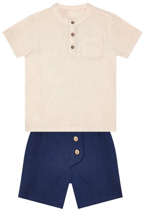 Camicia beige a maniche corte con tre bottoni sul davanti e taschino sul petto, abbinata a pantaloncini blu navy con due bottoni davanti.