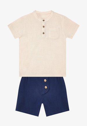 Camicia beige a maniche corte con tre bottoni sul davanti e taschino sul petto, abbinata a pantaloncini blu navy con due bottoni davanti.
