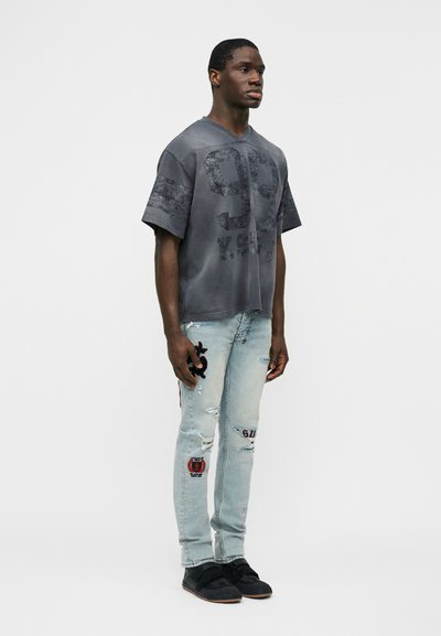 Άνδρας στέκεται στραμμένος ελαφρώς δεξιά, φορώντας φαρδιά σκουρόγριζα μπλούζα με τύπωμα "99", ανοιχτόχρωμο μπλε τζιν με σκισίματα και μπαλώματα, και μαύρα παπούτσια.