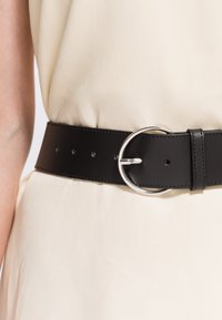 Zwarte leren riem met een ronde zilveren gesp, vastgemaakt rond de taille van een crèmekleurige jurk.
