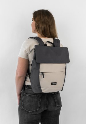 Zaino con un cappuccio superiore grigio e corpo color crema. Presenta spallacci regolabili, una tasca frontale con zip e un'etichetta con il logo del marchio.