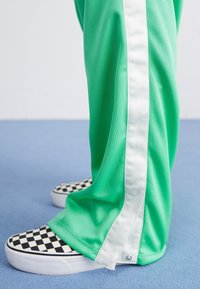 Green athletic pants with white side stripes, paired with black and white checkered slip-on shoes, textured fabric.