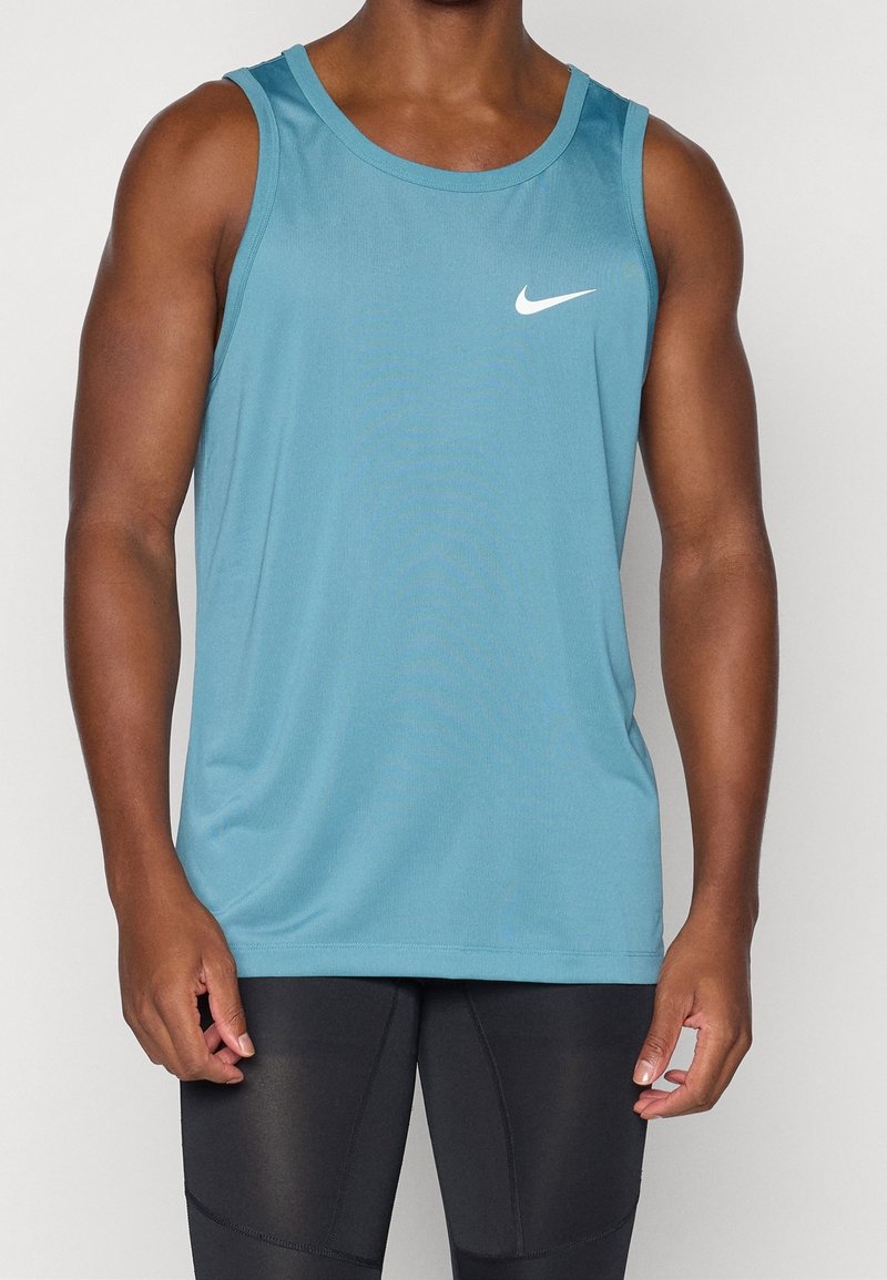 Homme portant un débardeur Nike sans manches turquoise et des leggings de sport noirs ajustés, debout devant un fond clair uni.