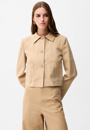 Frau trägt eine beige Jacke mit Knöpfen und zwei Brusttaschen sowie passende hoch taillierte Hose, steht vor weißem Hintergrund.
