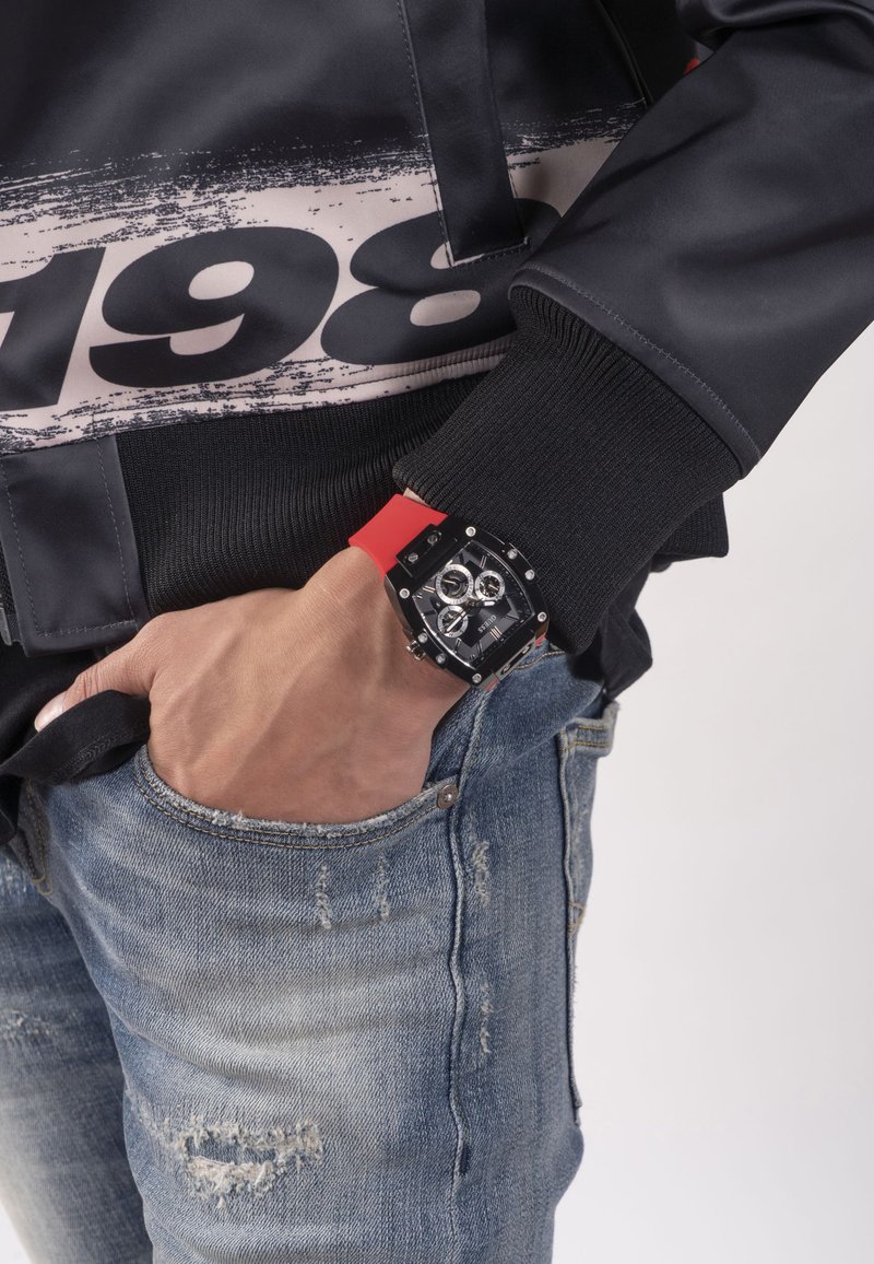 Schwarze und rote Chronographuhr mit quadratischem Ziffernblatt, Gummiband und silbernen Akzenten; getragen mit schwarzer Jacke und ausgewaschenen blauen Jeans.