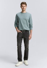 Šviesiai mėlyna ilgomis rankovėmis marškiniai, tamsiai pilkos spalvos džinsai, juodas diržas ir baltos sportinės avalynės; modelis stovi rankomis kišenėse prieš neutralią foną.