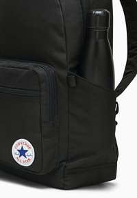 Mochila negra hecha de un tejido duradero, con un bolsillo lateral que contiene una botella de agua negra y un logo circular de Converse en la parte frontal.