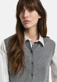 Gilet grigio lavorato a maglia con chiusura a bottoni, collo alto e bordi a costine, indossato sopra una camicia bianca a strisce verticali sottili.