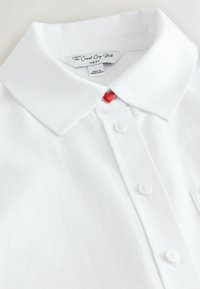 Bela kratka obrezana srajca s kratkimi rokavi, s koničastim ovratnikom, petimi belimi gumbi in rdečim gumbom v obliki srca na izrezu.