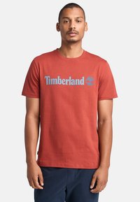 Červené bavlněné tričko s modrým logem "Timberland" a grafikou stromu. Okrúhly výstřih a krátké rukávy s rovnými lemy.
