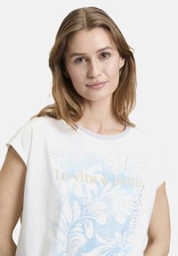 White short-sleeve T-shirt featuring a blue floral pattern and text "La vita è bella" in beige, accented with rhinestones.
