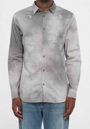 Hombre con una camisa gris de manga larga abotonada con patrones sutiles de "AX" y jeans azules, de pie contra un fondo claro.