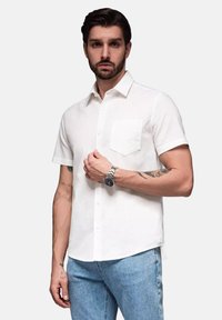 Witte kortermige overhemd met knoopsluiting en een enkele borstzak, gecombineerd met lichtblauwe jeans. Het model heeft tatoeages en draagt een zilveren horloge.