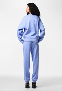 Femme aux cheveux bouclés debout, portant un sweat-shirt bleu clair et un pantalon de jogging assorti avec des baskets noires, de dos sur un fond uni.
