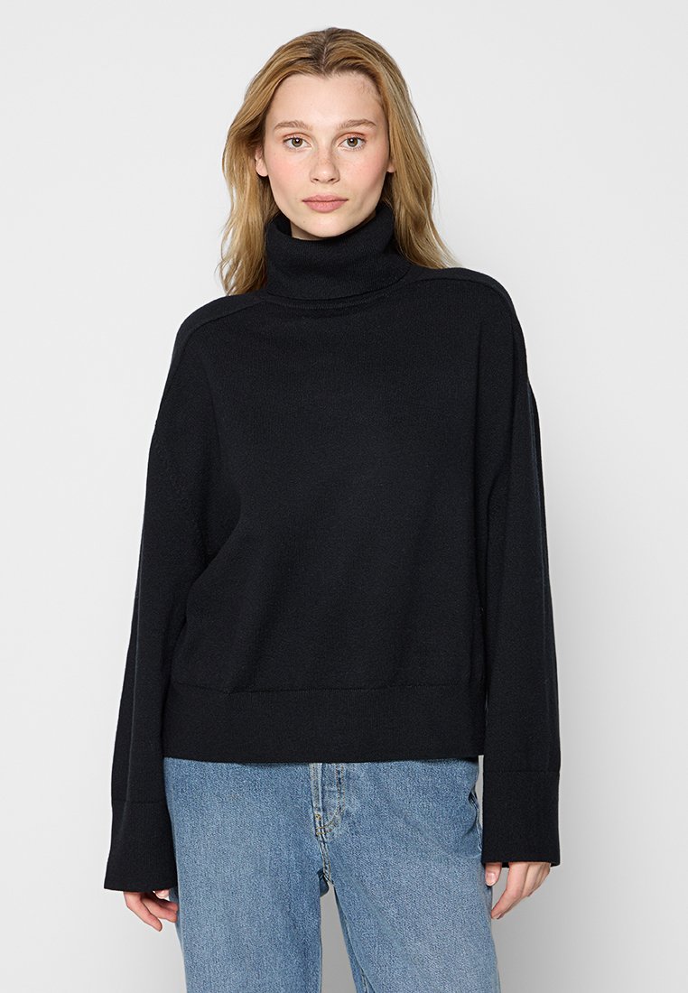 Młoda kobieta w czarnym swetrze z golfem i niebieskich dżinsach, stojąca na tle jasnoszarej ściany.