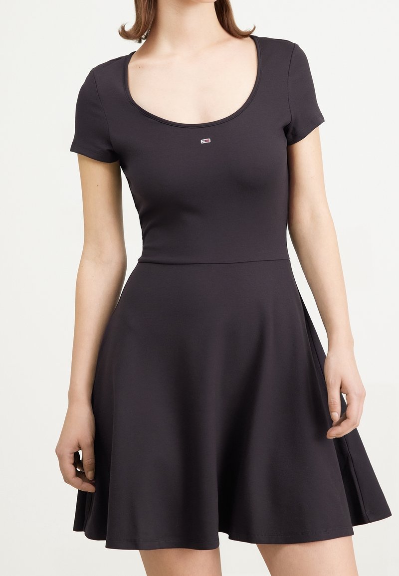 Schwarzes, kurzärmeliges Kleid mit Rundhalsausschnitt, aus weichem, elastischem Stoff. Es hat ein tailliertes Oberteil und einen schwingenden Rock, mit einem kleinen Logo-Akzent.
