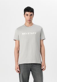 Γκρι βαμβακερό μπλουζάκι με κοντά μανίκια και στρογγυλή λαιμόκοψη. Διαθέτει λευκή επιγραφή "BELSTAFF" στο στήθος. Συνδυάζεται με σκούρες γκρι τζιν.