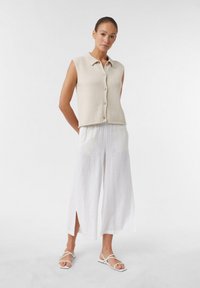 Beżowy bezrękawnik z wąskim kołnierzykiem i guzikami, zestawiony z białymi szerokimi spodniami lnianymi z bocznymi rozcięciami, uzupełniony o sandały na paski.