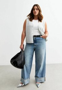 Lichtgrijze mouwloze cardigan over een witte top, gecombineerd met brede blauwe jeans. Een zwarte handtas en zilveren puntenflatjes maken de look compleet.