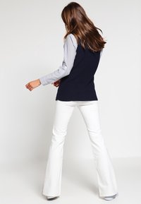 Námořnický žebrovaný bezrukávový svetr přes světle modrou pruhovanou košili s dlouhým rukávem, spárovaný s bílými zvonovými kalhotami. Hladká tkanina a casual střih.