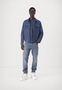 Τζάκετ τζην σε μπλε χρώμα, από βαμβακερό ύφασμα, με δύο τσέπες στο στήθος. Φορεμένο πάνω από ένα μπλε φούτερ και συνδυασμένο με ανοιχτό μπλε τζιν και λευκά αθλητικά παπούτσια.