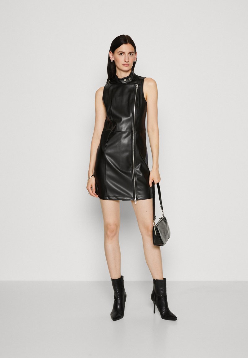 Abito senza maniche in pelle nera con colletto alto, zip frontale e silhouette aderente. Abbinato a stivali neri alla caviglia e una borsa piccola.