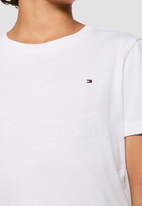 Camiseta blanca de algodón con cuello redondo, mangas cortas y un pequeño logo en rojo, blanco y azul en el pecho. Textura suave.