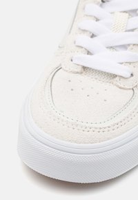 Tenisky s měkkým nubukovým svrškem v světlé béžové barvě, s bílými tkaničkami, jemnými perforacemi a texturovanou bílou gumovou podrážkou.