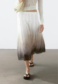 Jupe midi plissée avec un effet ombragé transitionnant du crème clair au gris foncé, fabriquée en tissu léger. Portée avec des tongs noires.