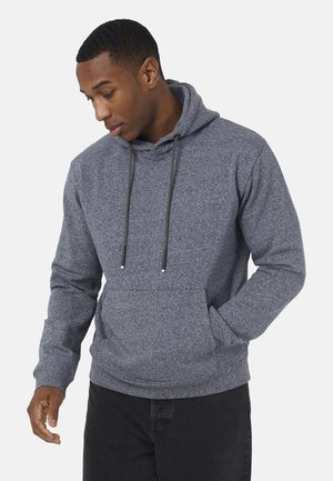 Man draagt een grijs gemêleerde hoodie met zwarte trekkoorden, één hand in de zak, kijkt naar beneden en iets opzij.