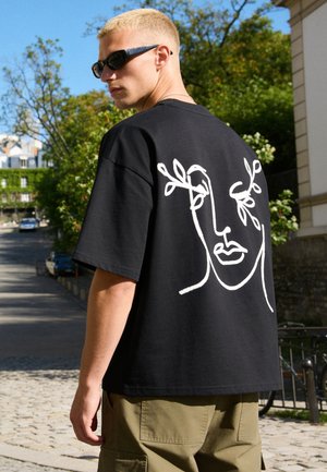 Junger Mann mit kurzen blonden Haaren, der schwarze Sonnenbrillen und ein schwarzes T-Shirt mit einer weißen abstrakten Gesichtzeichnung auf dem Rücken im Freien trägt.